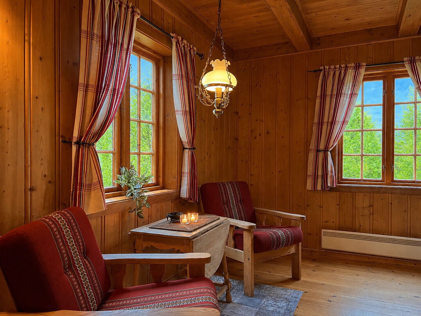 A wooden interior with two windows, a hanging light fixture, a table with candles, and two chairs.