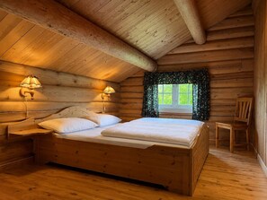 Un dormitori d'una cabana de fusta amb un llit, dues làmpades murals, una finestra amb cortines i una cadira.