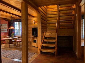 Interior d'una cabana de fusta amb zona de menjador, televisió i escala cap a una zona alta.