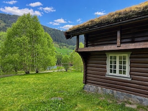 A wooden cabin with a stone foundation, surrounded by lush green grass and trees, set against a backdrop of mountains and a clear sky.