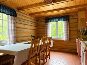A wooden cabin interior with a dining table, chairs, a window with curtains, and a kitchen area with cabinets and utensils.