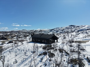 A cabin with a dark roof, surrounded by snow-covered terrain and sparse trees. - New spacious mountain cabin with sauna, Geilo (Hol)
