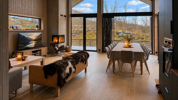 A modern interior with a fireplace, dining area, and large windows offering a view of the outdoors. - New spacious mountain cabin with sauna, Geilo (Hol)