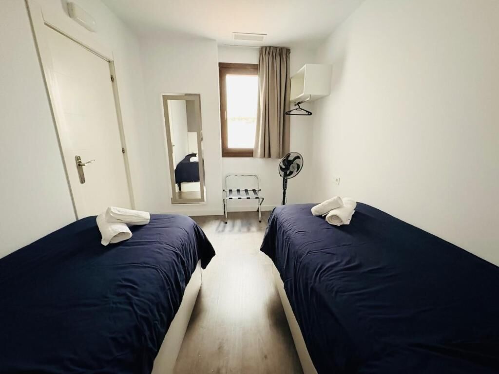 A hotel room with two single beds, a bedside table, a mirror, a window with curtains, a standing fan, and a door.