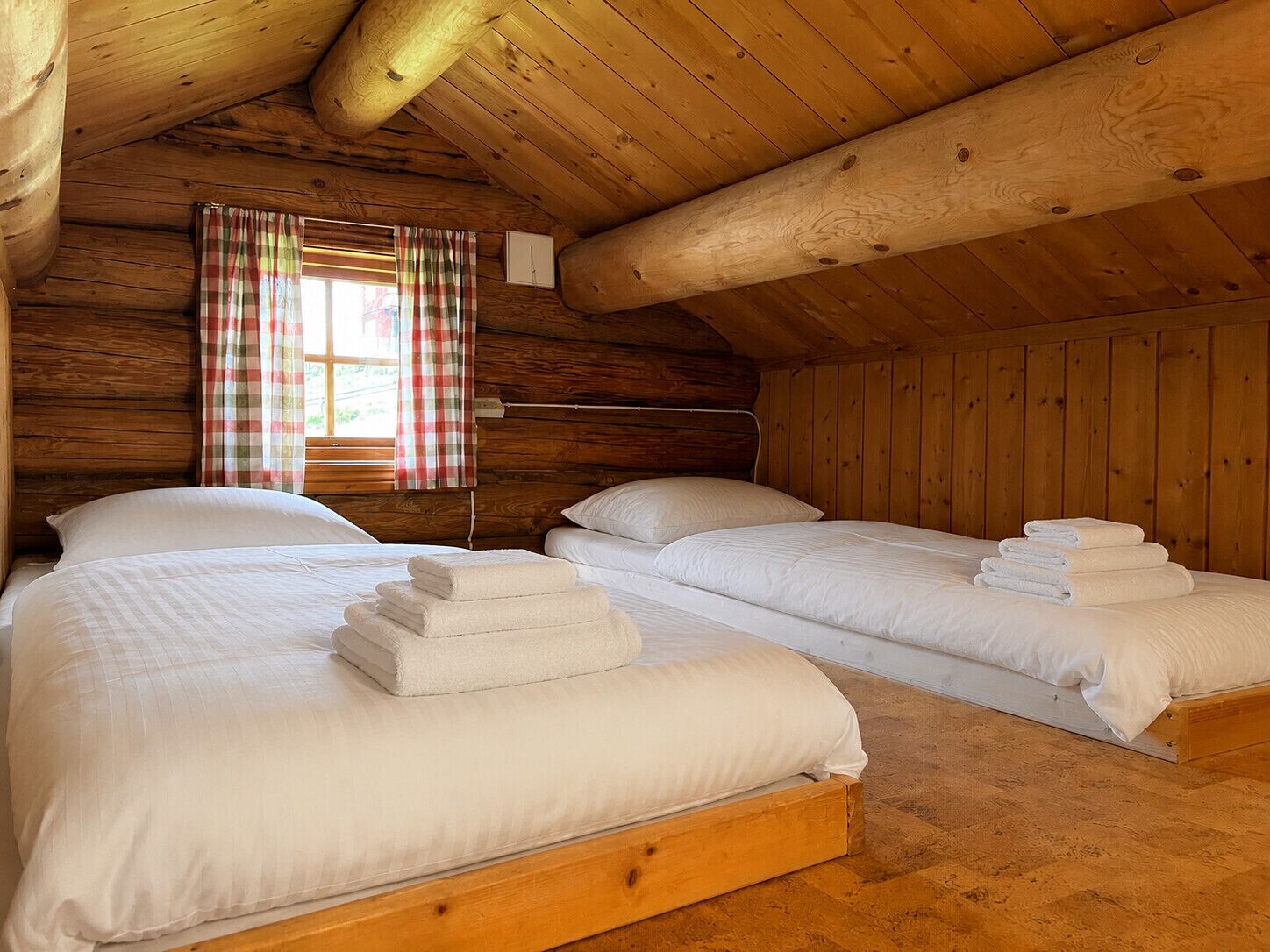 A wooden cabin room with two beds, a window with plaid curtains, and a wooden ceiling.