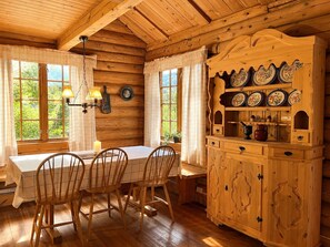A wooden cabin interior with a dining table, chairs, a cabinet with plates, and a window with curtains.