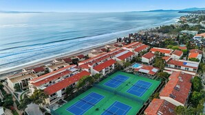 Un hotel costero con varias canchas de tenis, vistas a la playa y una zona residencial densa al fondo.