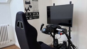 Una silla gamer con un monitor y pedales, un radiador y un cartel en la pared.
