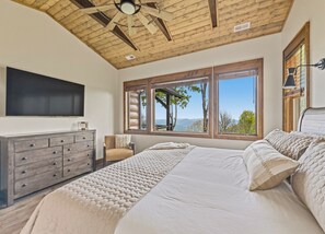 A bedroom with a bed, a dresser, a TV, and a window with a view of trees and mountains. - Gold Rain Retreat Newly Built! 4BD,3.5 BA in Smoky Mountain Retreat (Maggie Valley)