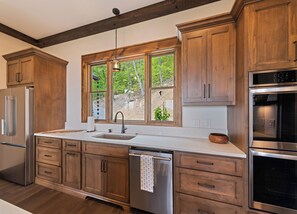 A kitchen with wooden cabinets, a stainless steel refrigerator, a dishwasher, and a large window with a view of greenery. - Gold Rain Retreat Newly Built! 4BD,3.5 BA in Smoky Mountain Retreat (Maggie Valley)