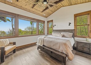 A bedroom with a large bed, wooden beams, multiple windows, and a view of trees. - Gold Rain Retreat Newly Built! 4BD,3.5 BA in Smoky Mountain Retreat (Maggie Valley)