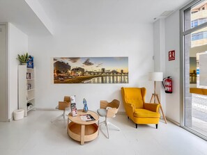 A modern interior with a cityscape painting, a round wooden table, a yellow armchair, and a bookshelf. - Guestready - Nuevos Livian Suites Las Palmas (Las Palmas de Gran Canaria)