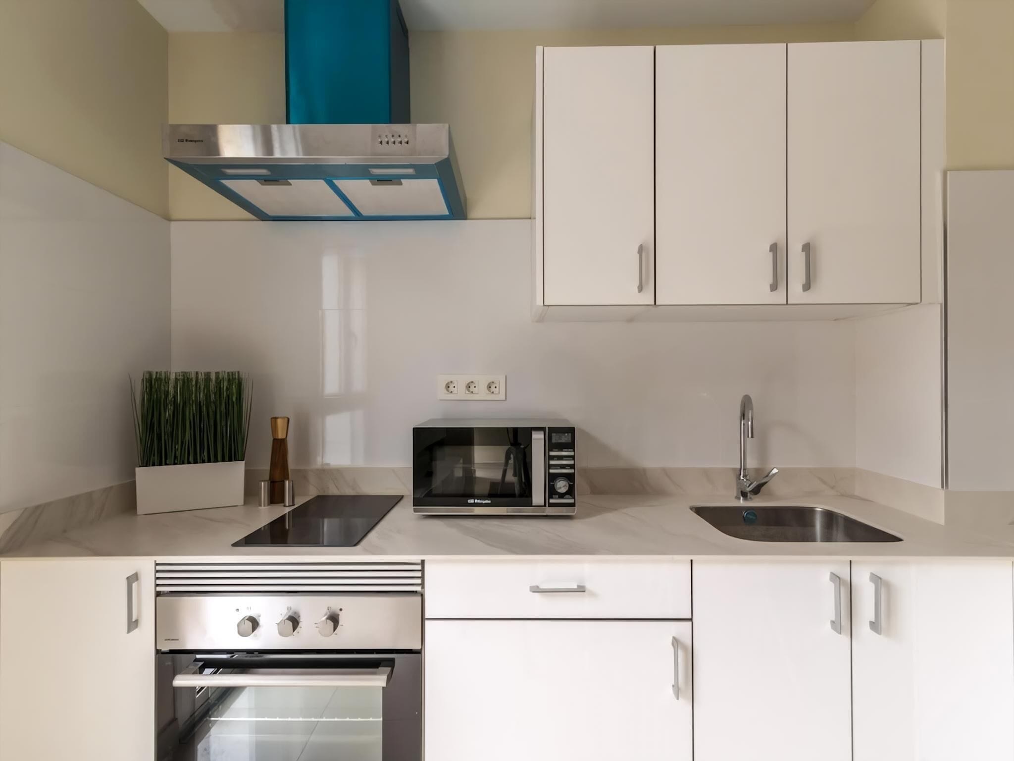 Una cocina moderna con armarios blancos, horno empotrado, microondas, lavaplatos y una campana extractora azul.