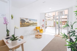A hotel lobby with a reception desk, chairs, a large window with a cityscape view, and a door leading outside. - Guestready - Nuevos Livian Suites Las Palmas (Las Palmas de Gran Canaria)