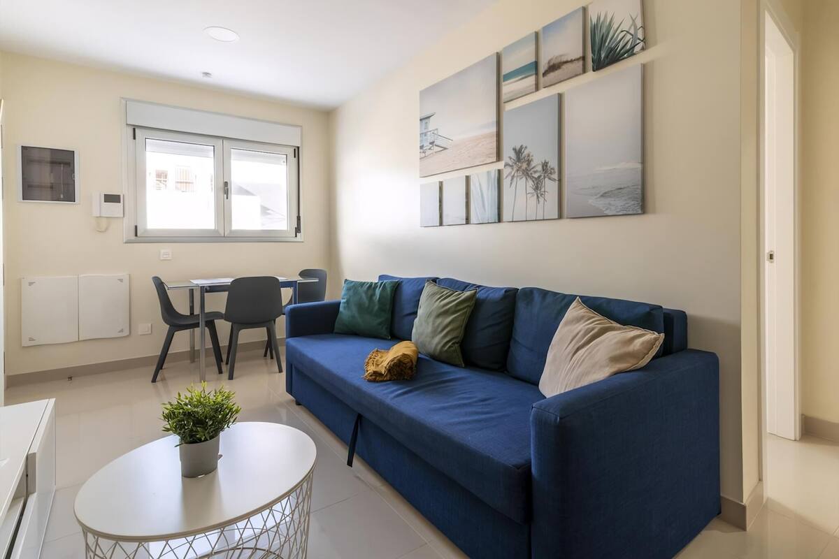 A modern living room with a blue sofa, a small round table with a plant, a dining area with a table and chairs, and a window with blinds.