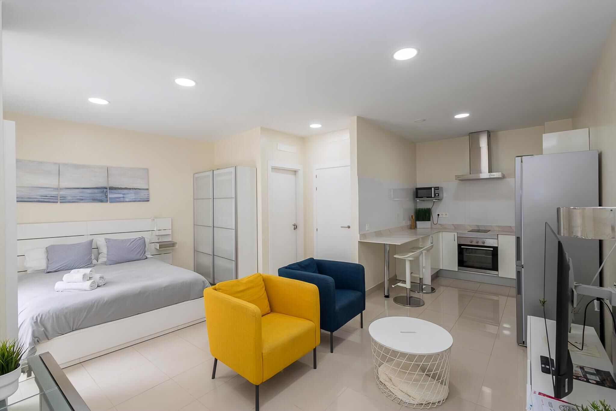 A modern hotel room with a bed, two chairs, a small table, a kitchen area with a microwave, stove, and refrigerator.