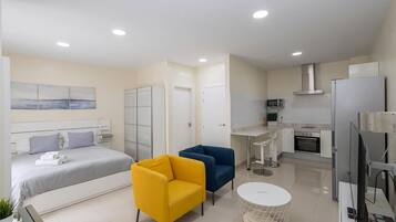 A modern hotel room with a bed, two chairs, a small table, a kitchen area with a microwave, stove, and refrigerator.