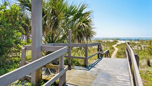 Ein hölzerner Steg führt zu einem Strand mit Palmen und blauem Himmel.