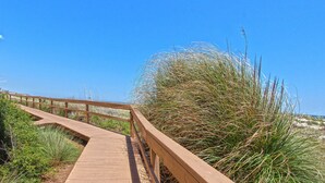 Ein hölzerner Steg führt zu einem Strand mit hohem Gras und wolkenlosem, blauem Himmel.
