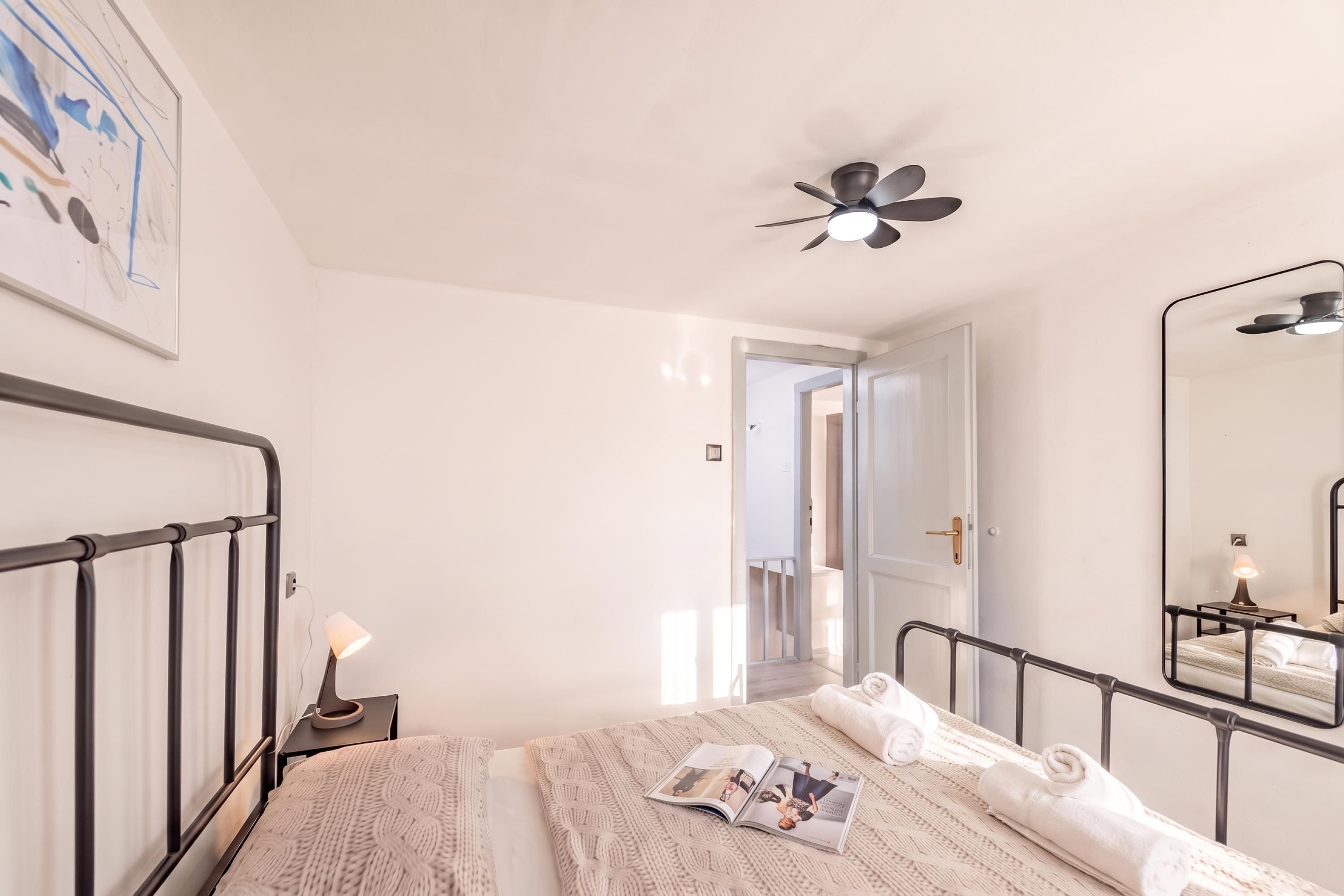 Ein Schlafzimmer mit einem Bett, einer Nachttischlampe, einem Deckenventilator, einem Spiegel und einer Tür zu einem anderen Raum.
