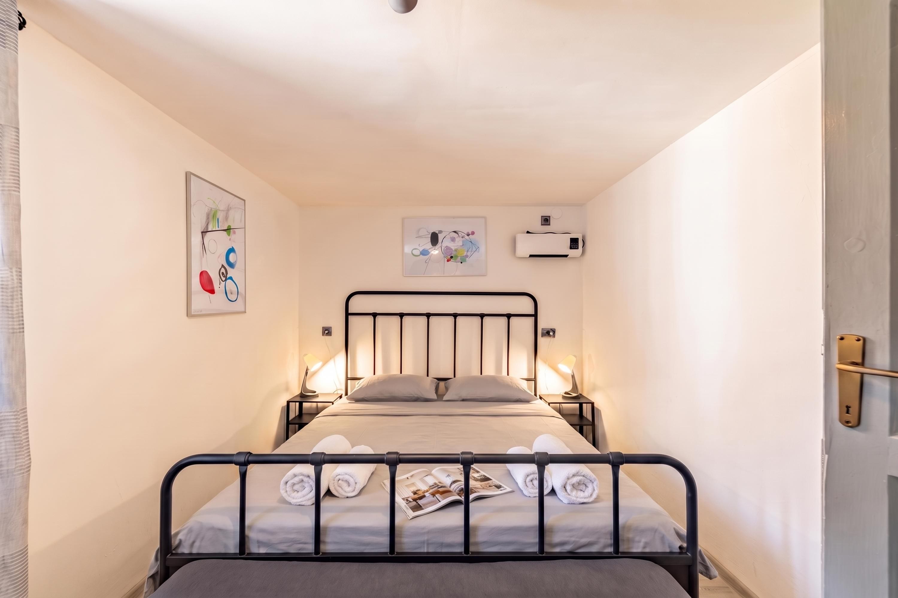 A single bed with a metal frame, two bedside lamps, a wall-mounted air conditioner, and two framed artworks.