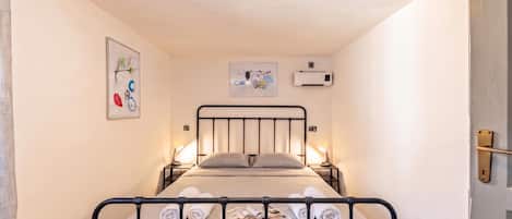 A single bed with a metal frame, two bedside lamps, a wall-mounted air conditioner, and two framed artworks.