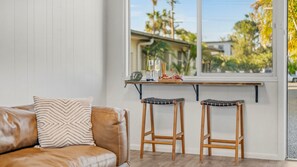 A room with a white wall, a brown leather sofa, two wicker stools, a window with a view of palm trees, and a small table with a bottle, glasses, and a fruit platter.