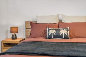 Ein Bett mit dunkler Decke, zwei Kissen mit Palmenmotiven, ein Nachttisch aus Holz mit einer Lampe und ein Kopfteil.