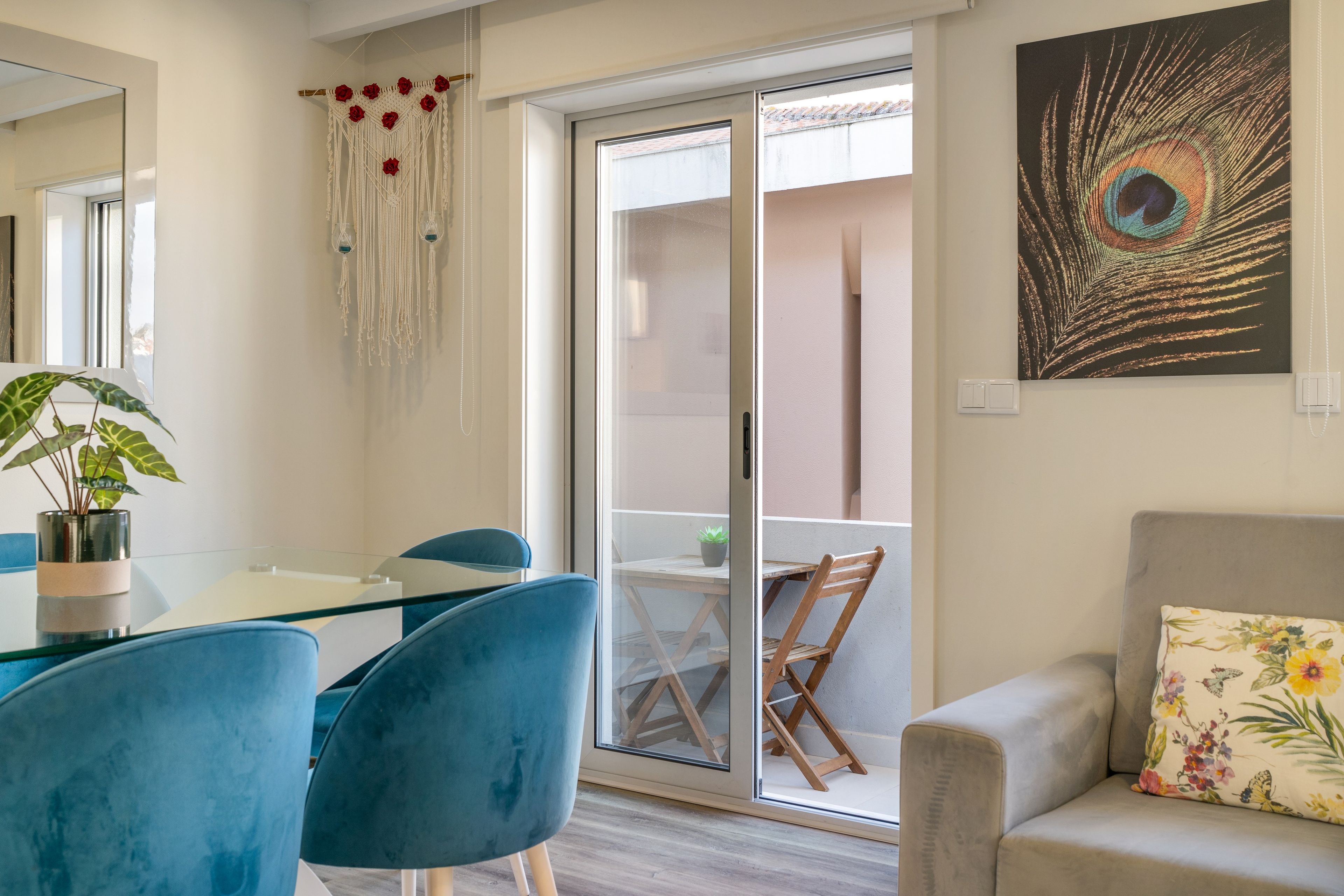 Un salon moderne avec une table à manger en verre, des chaises bleues, un canapé gris et une œuvre d’art représentant des plumes de paon.