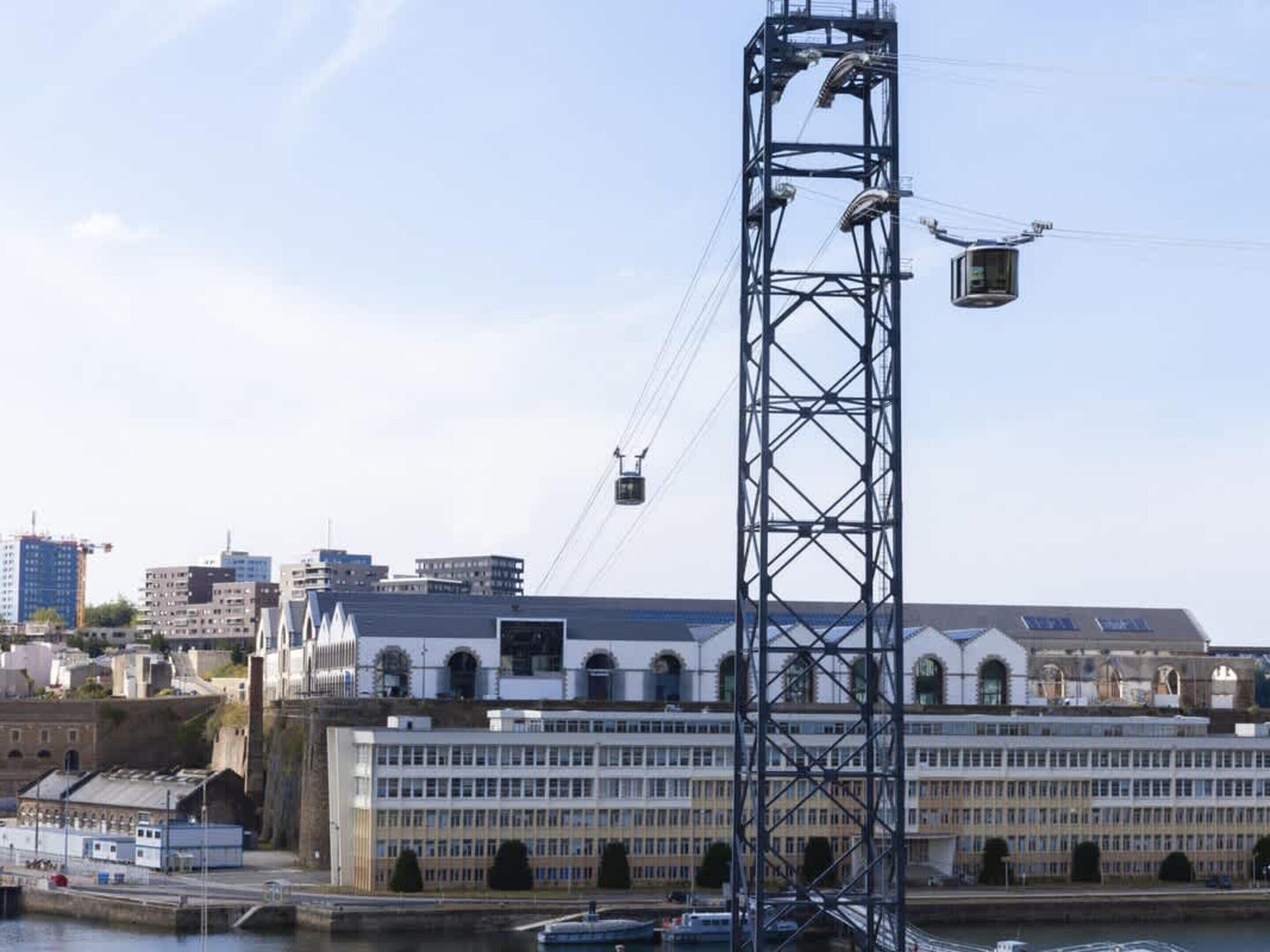 A tall metal structure with a cable car system, a large building with arched windows, and a body of water with boats.