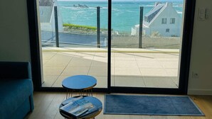 A room with a large glass door leading to a balcony with a view of the sea and buildings. - Apartment in Porspoder Near Plage du Bourg (Porspoder)