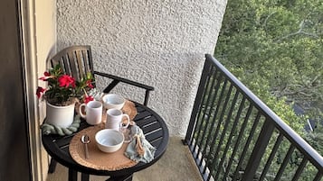 Een balkon met een tafel gedekt voor twee personen, een stoel, een bloempot en uitzicht op bomen.