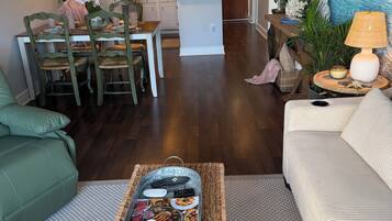 Un soggiorno con divano, tavolino da caffè con riviste e telecomandi, zona pranzo con sedie e tavolo, e una cucina sullo sfondo.