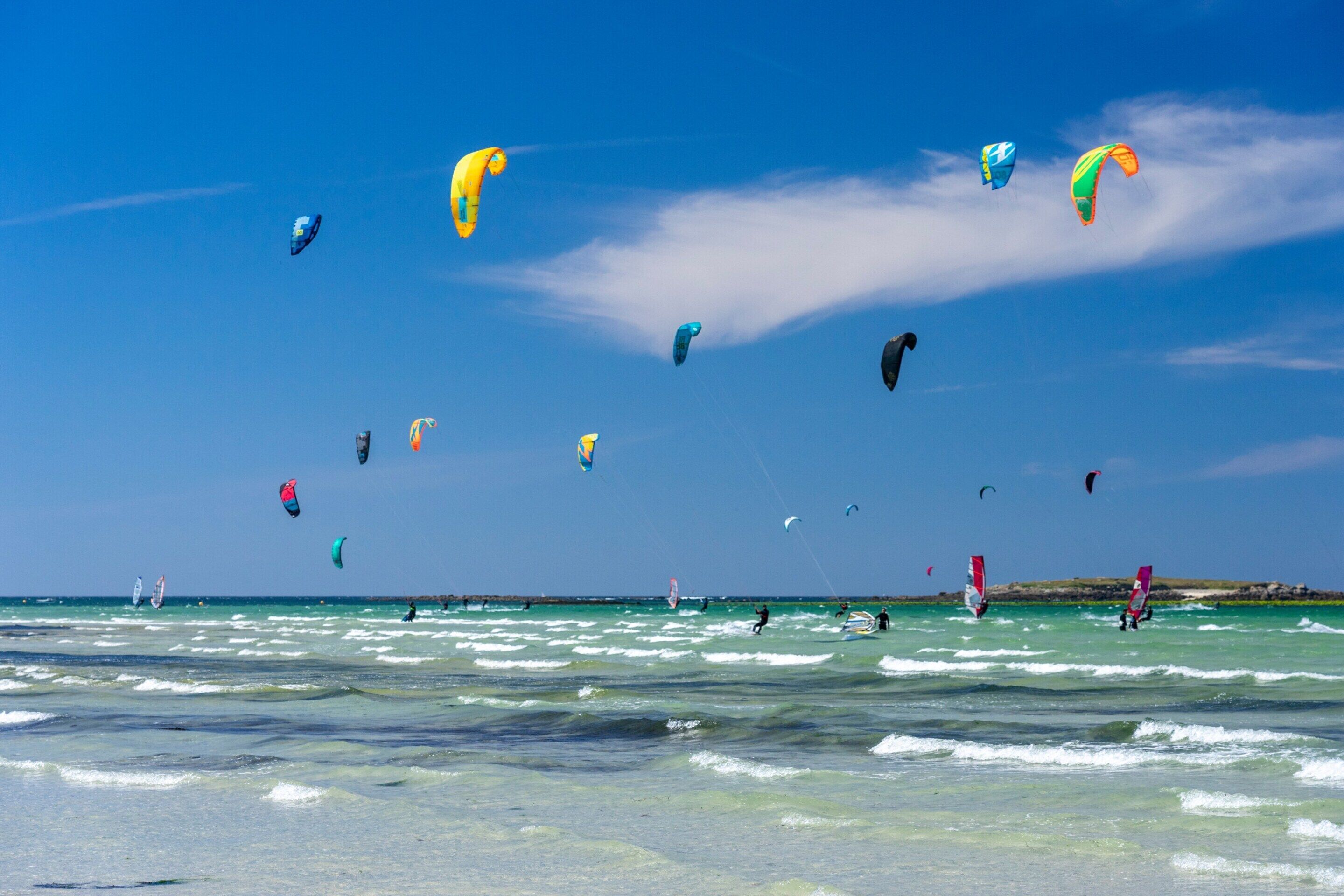 A beach with multiple kites in the sky and people windsurfing.