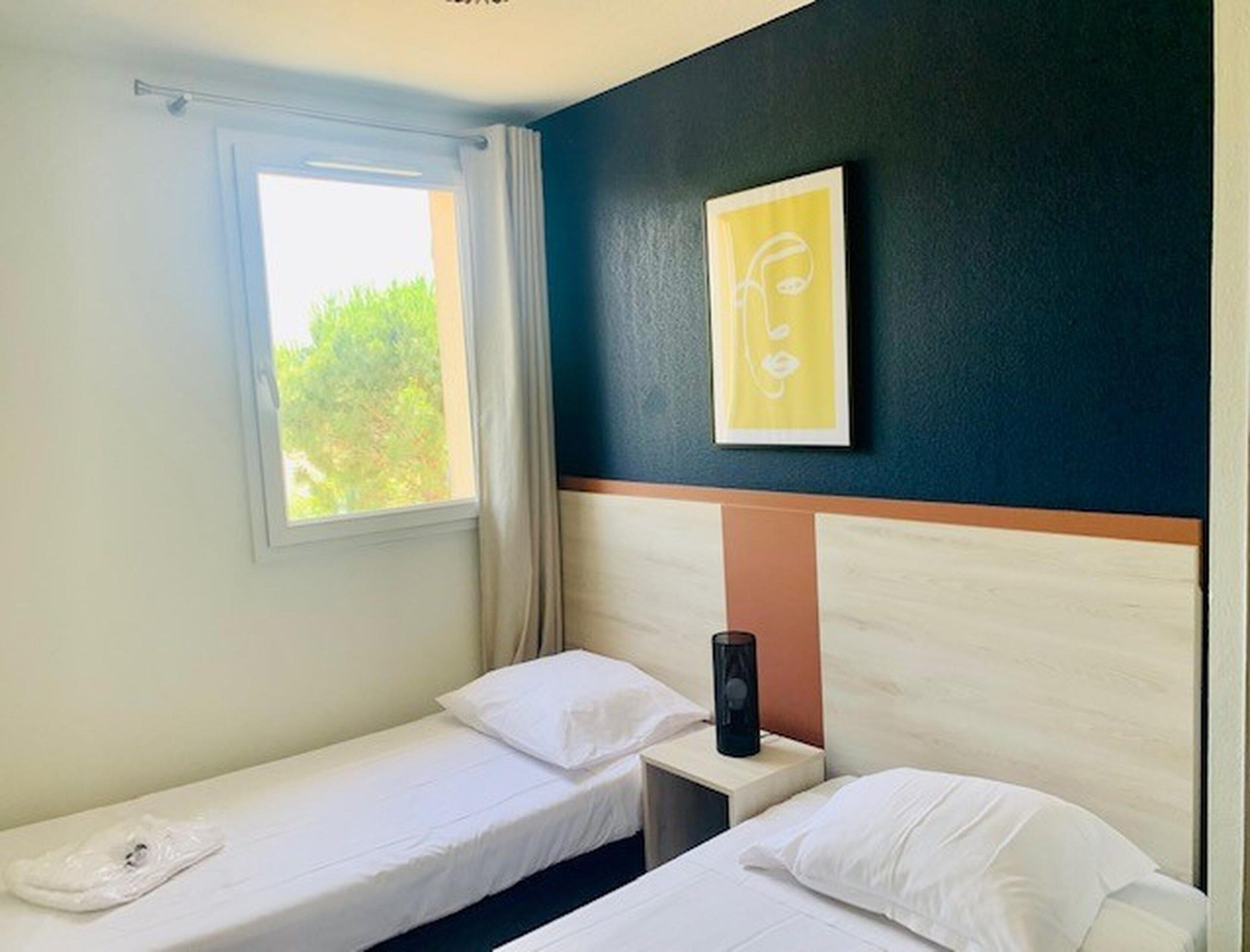 A hotel room with two single beds, a window with curtains, a bedside table with a lamp, and a wall-mounted artwork.
