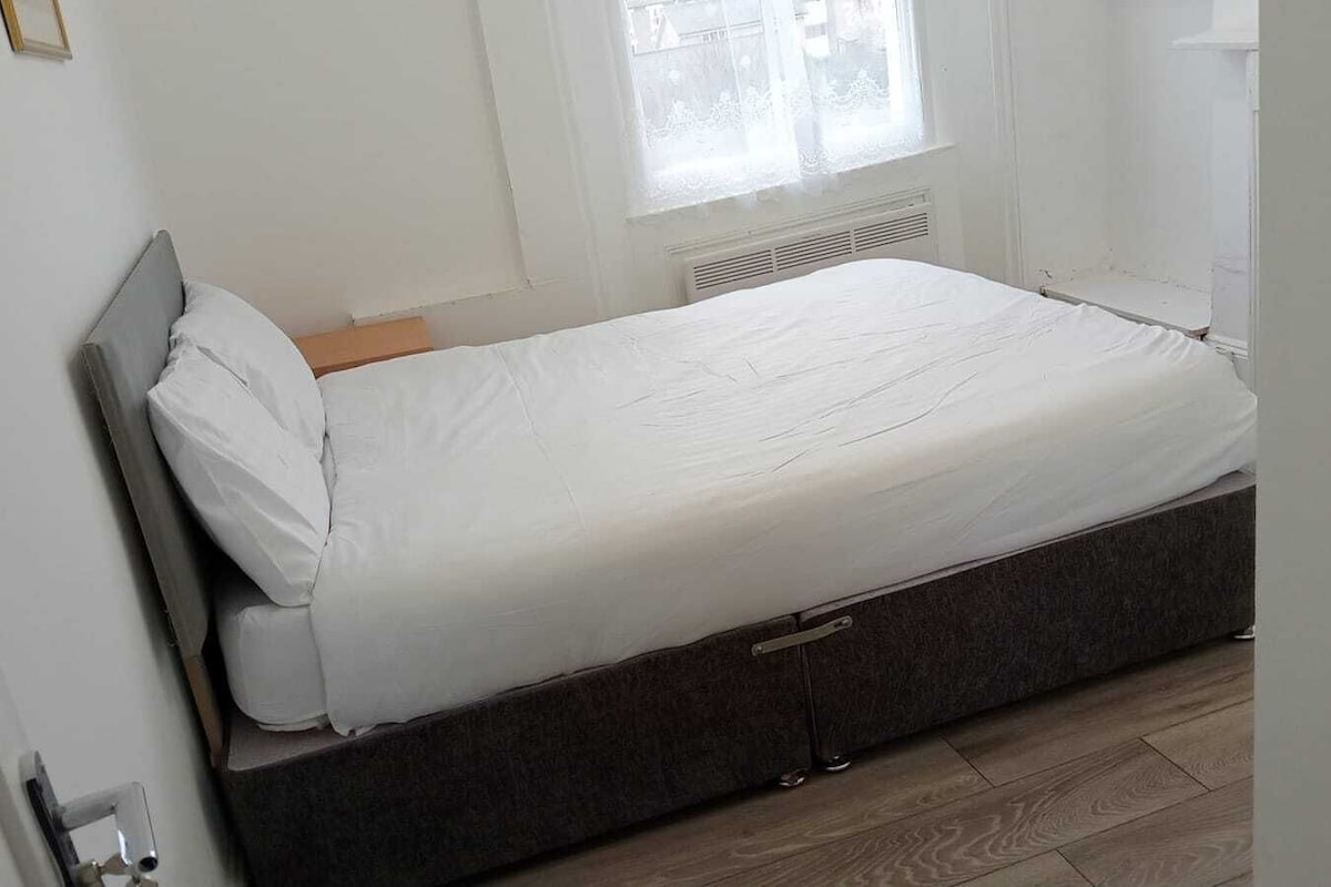 Ein Einzelbett mit dunklem Kopfteil in einem Zimmer mit weißen Wänden und einem Fenster mit durchsichtigen Vorhängen.
