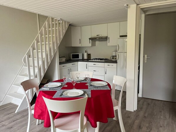 Un coin repas avec une nappe rouge, des chaises blanches et une cuisine en arrière-plan.