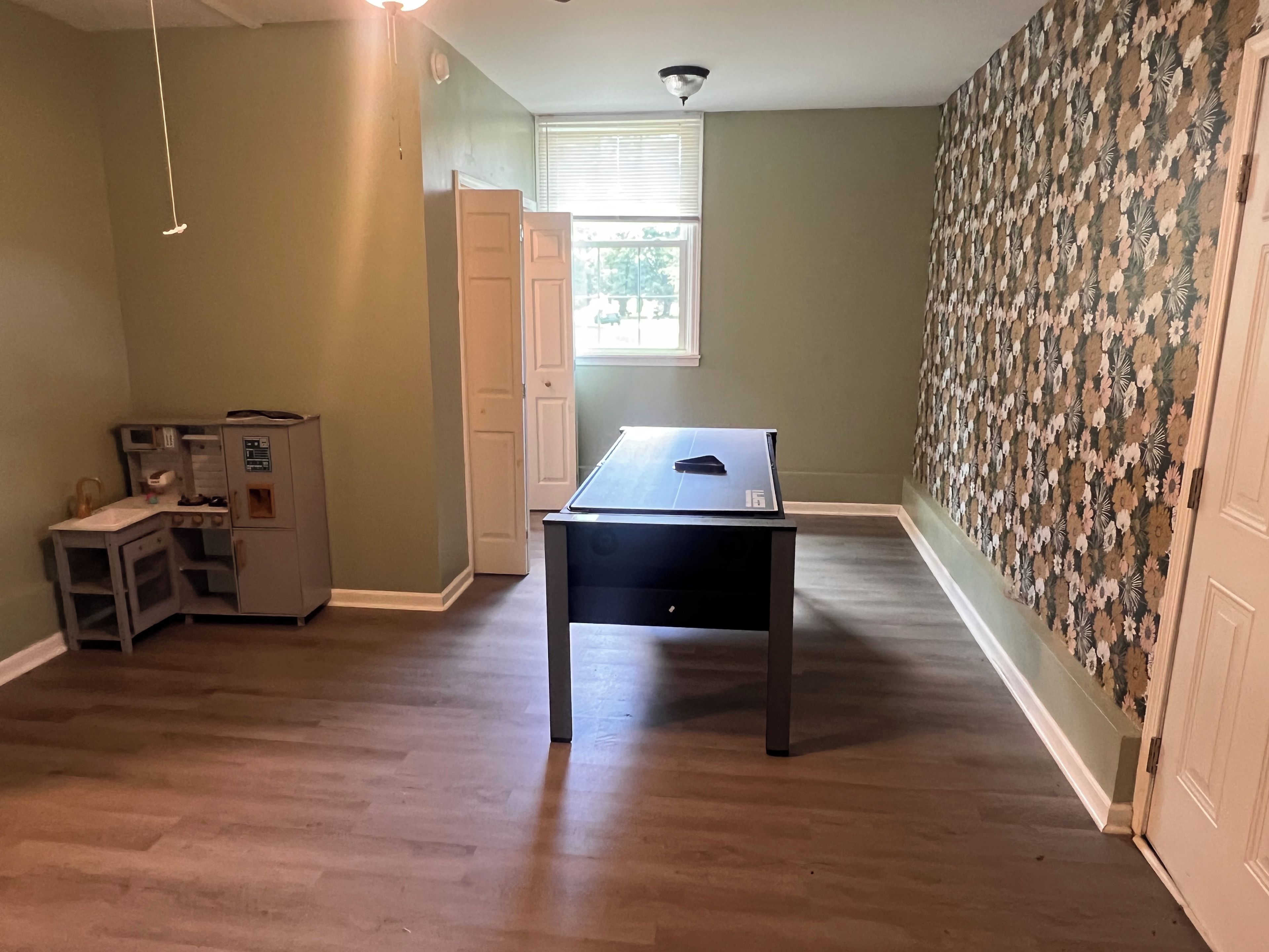 A room with a wooden floor, a table, a play kitchen, and a floral wallpaper.