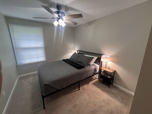 A bedroom with a bed, a nightstand, a lamp, a ceiling fan, and a window with blinds.