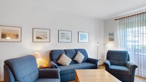 Ein Wohnzimmer mit Sofa, Sessel und Couchtisch, gerahmten Bildern an der Wand und einem Fenster mit Vorhängen.