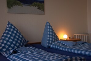Een slaapkamer met een geruit bedovertrek, kussens, een nachtkastje met een lamp en een schilderij aan de muur.