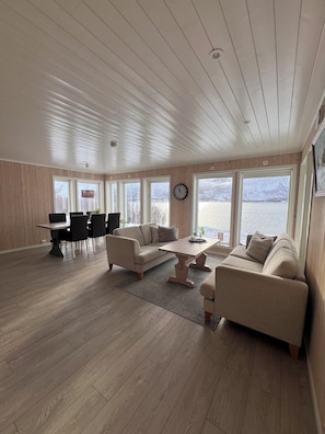 Amplia sala de estar con piso de madera, un conjunto de sofás, una mesa de comedor y ventanas grandes que ofrecen vistas al exterior.