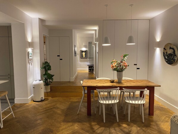 Ein moderner Essbereich mit einem Holztisch, weißen Stühlen, Pendelleuchten und einem Blumenstrauß.