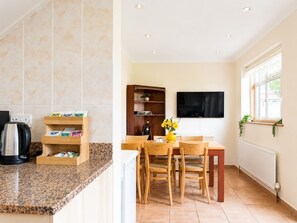 A kitchen with a granite countertop, a coffee maker, a shelf with tea boxes, a dining table with chairs, a TV mounted on the wall, and a window with plants. - Ashwood Stays
Where Comfort Meets Elegance,Your Comfortable Home Away From Home (Chessington)