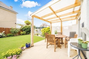 Une terrasse couverte avec une table et des chaises en bois, entourée d’un jardin et d’un mur blanc.