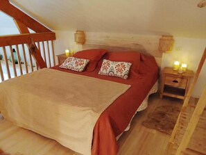 A bedroom with a wooden ceiling, a bed with red bedding and patterned pillows, two bedside tables with candles, and a wooden ladder.