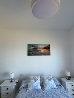 Un dormitorio con cama, dos mesitas de noche con lámparas, un cuadro colgado en la pared que muestra una escena de playa y una lámpara de techo blanca.