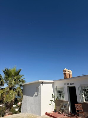 A white building with a chimney, a door, and a window with bars. There are two chairs outside and a palm tree in the background. - A&N Playa Madrid 1, Almayate (Vélez Málaga)
