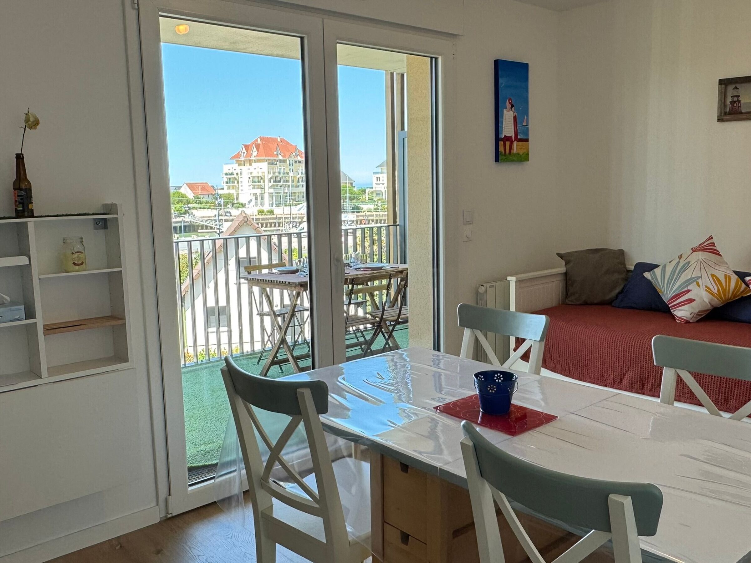 Una habitación con balcón, una mesa con sillas, una cama con almohadas y un cuadro en la pared.