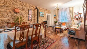 Ein gemütliches Wohnzimmer mit Steinwand, Essbereich mit Holzsesseln und Tisch, einer Couch, einem kleinen Tisch mit Lampe und einem Fenster mit Vorhängen.